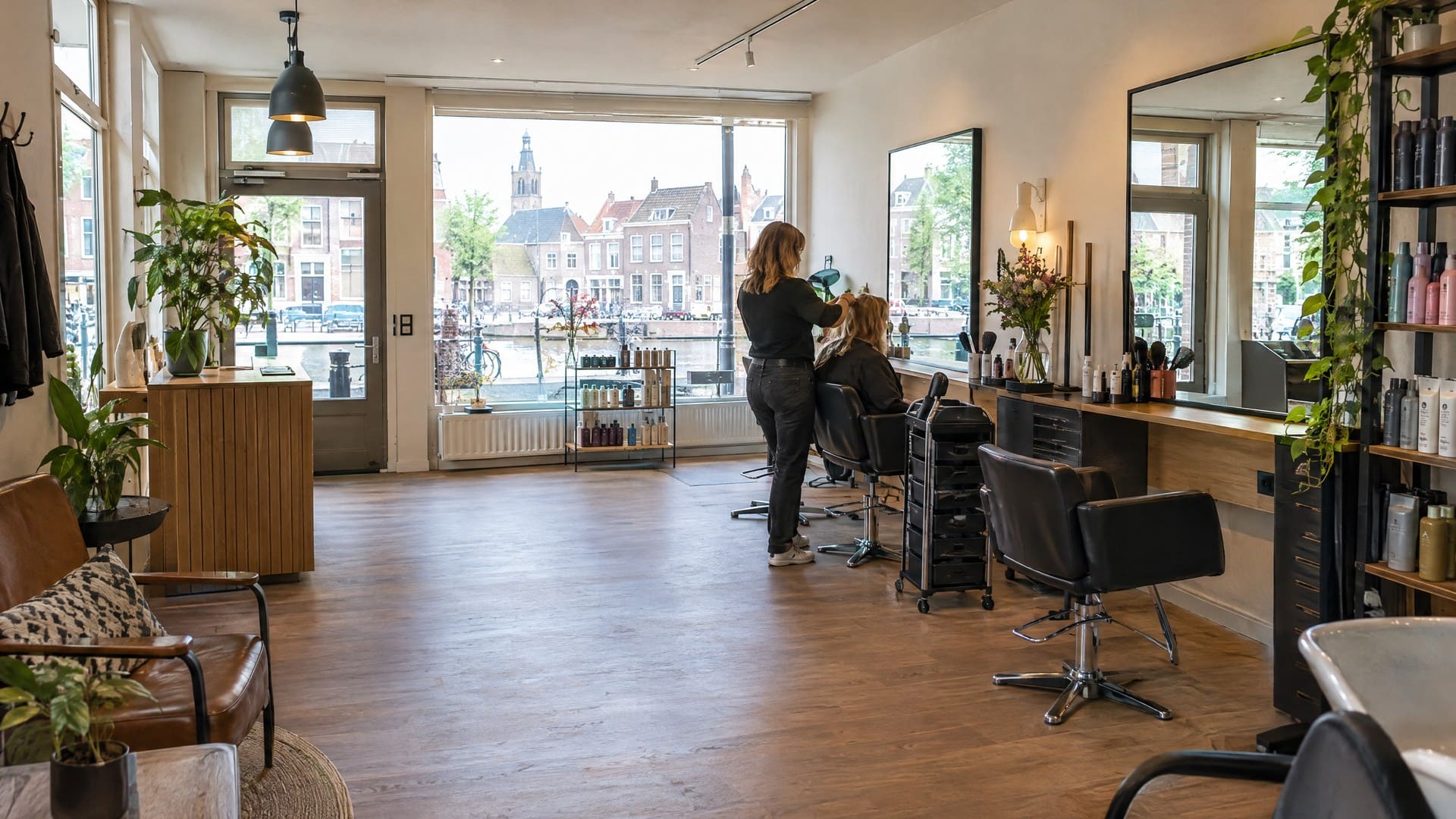 Hoek van een kapsalon met planten voor het raam, een houten balie en zicht op de straat buiten.