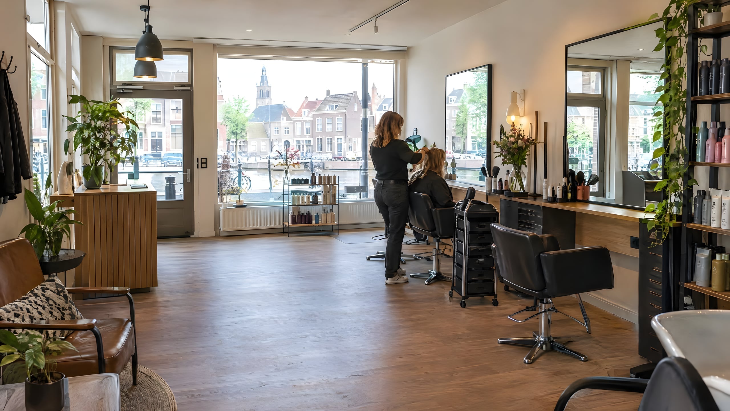 Lichte kapsalon met een houten vloer, grote ramen en uitzicht op een historische straat met oude huizen.